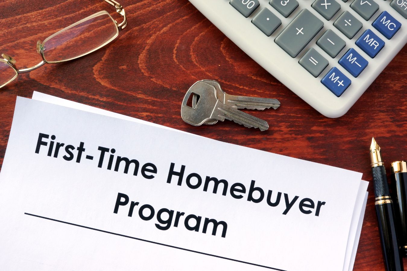 Keys, Eyeglass, Calculator and pen on a Brown Table with White Papers reading First Time Home Buyer Program