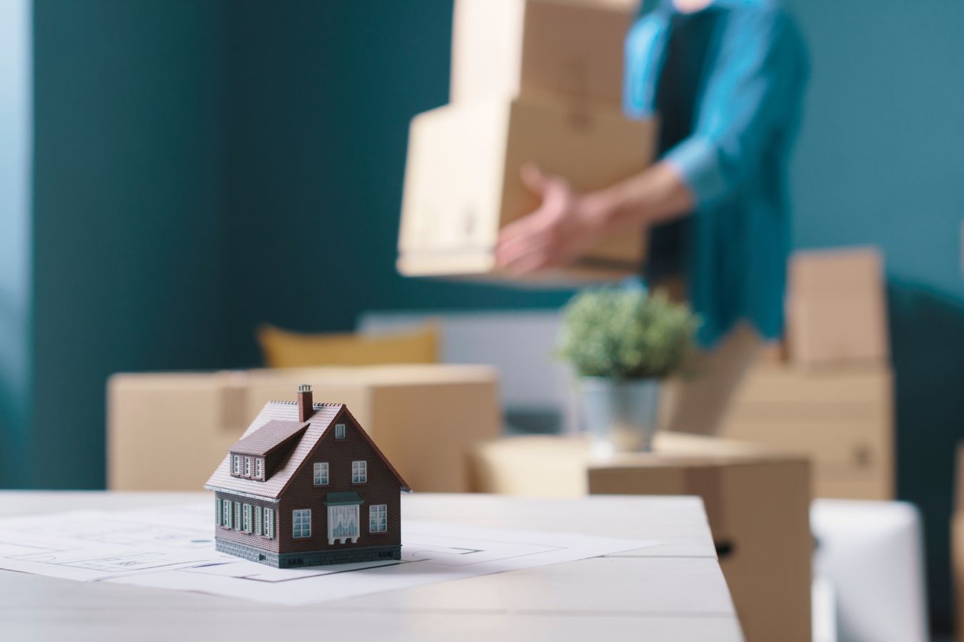 A man carrying boxes in hand and in front of him is a toy house on a table
