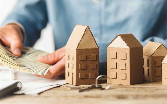 A hand counting cash next to model houses, symbolizing the potential for generating rental income through property investments